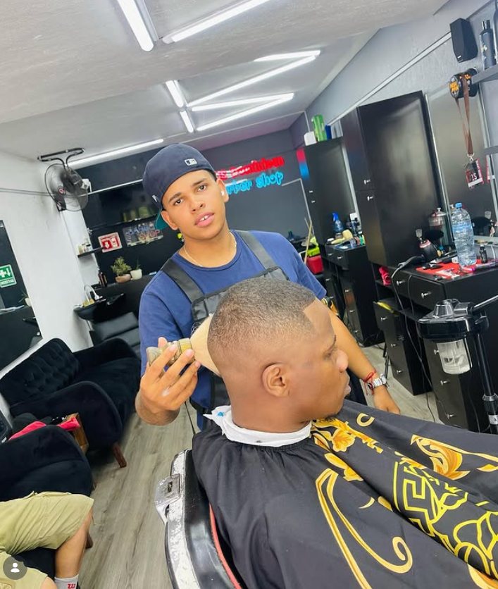 Barbero con máquina en mano mirando a la cámara junto a un cliente, con el letrero de neón de la sucursal al fondo