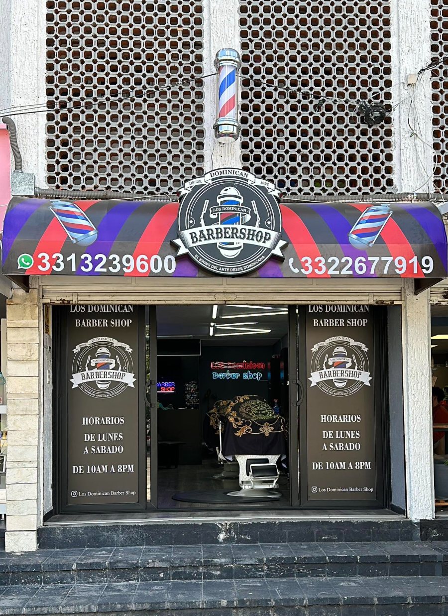 Fachada de Los Dominican Barbershop con su poste de barbero, toldo a rayas y logo sobre la entrada