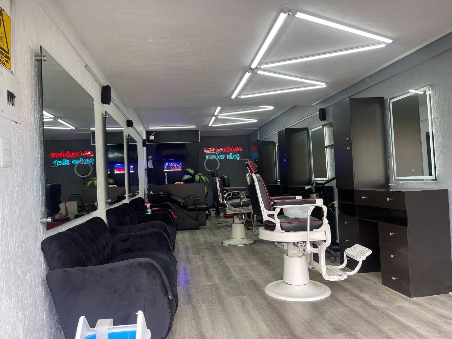 Vista amplia del interior de Los Dominican Barbershop con sillas blancas, espejos y letreros de neón al fondo